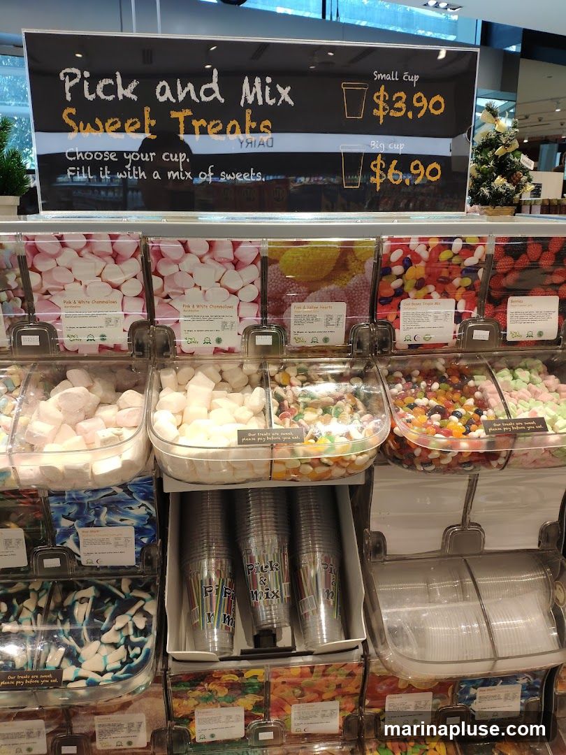 A display of various candies in bins, with cups for customers to fill. A sign above advertises "Pick and Mix Sweet Treats" with prices for small and big cups.
