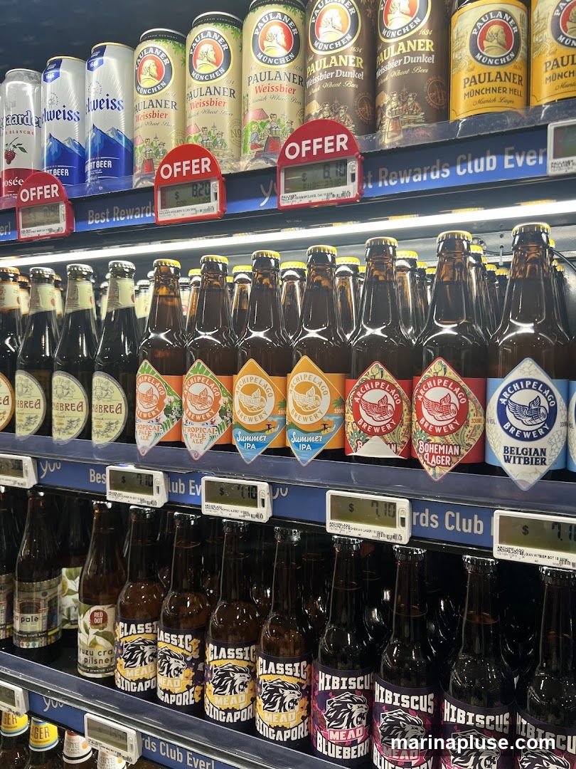 A shelf display of various beers and meads in cans and bottles. Brands visible include Paulaner, Archipelago, and various local Singaporean brands. Some items are marked with "OFFER" signs and prices.