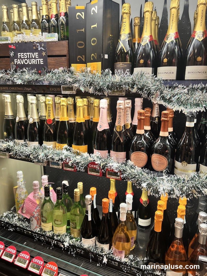 A display of various bottles of champagne and sparkling wine, with festive decorations. The bottles are arranged on shelves, with some bottles in boxes. A sign reads "FESTIVE FAVOURITE". Several price tags are visible, with prices such as $32.90, $33.50, and $14.00.