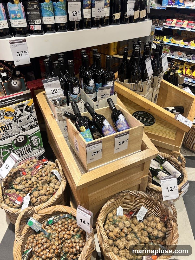A display of various bottles of wine and baskets of nuts in a retail setting. The wine bottles are arranged in wooden crates, with some priced at 29.00 and 30.00. Baskets of nuts, including hazelnuts and walnuts, are placed in front of the wine display, with prices ranging from 12.75 to 14.55. A sign for "Festive Favourite" is visible, along with a price of 37.00 for a bottle of wine.