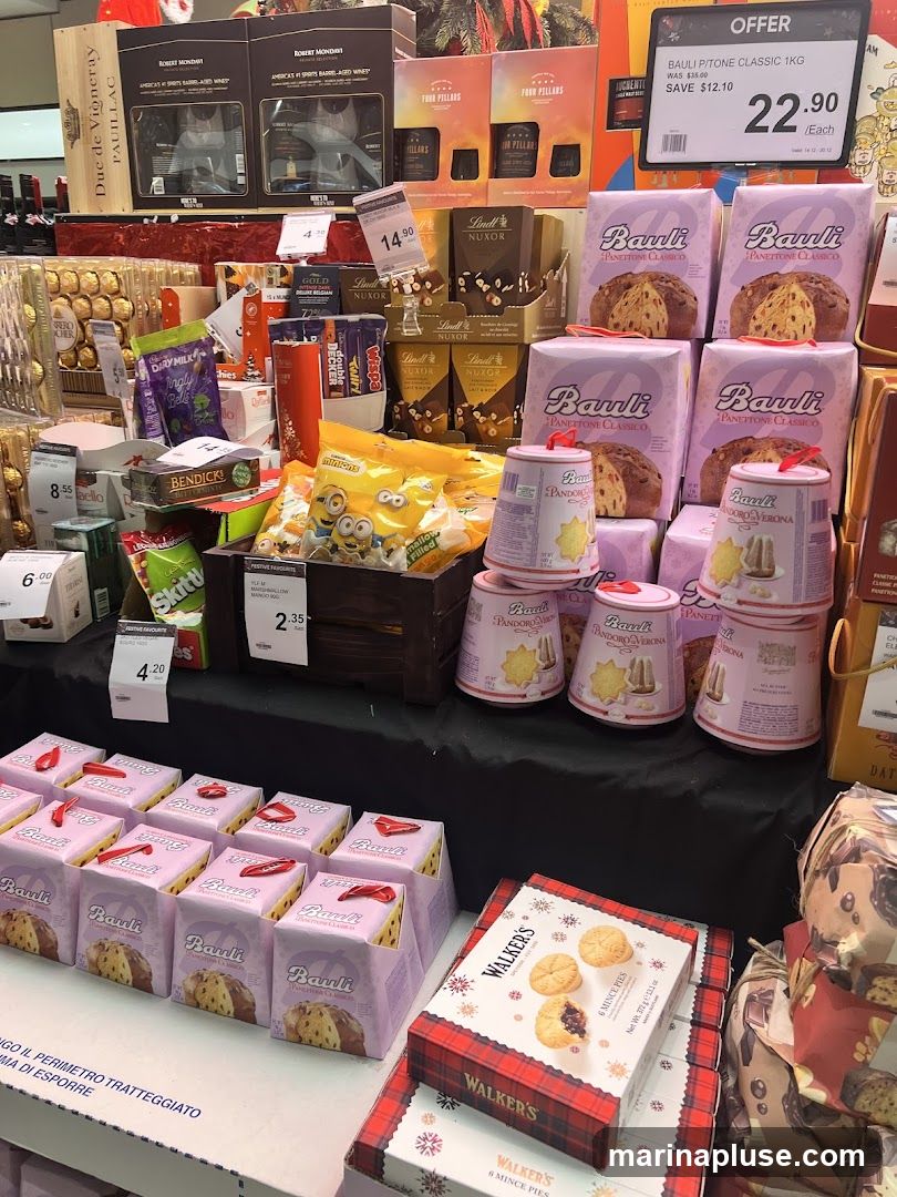 A display of various Christmas food items, including panettone, mince pies, chocolates, and wine. The display features products from brands like Bauli, Lindt, Walker's, and Robert Mondavi. Some items are on sale, with price tags indicating discounts.