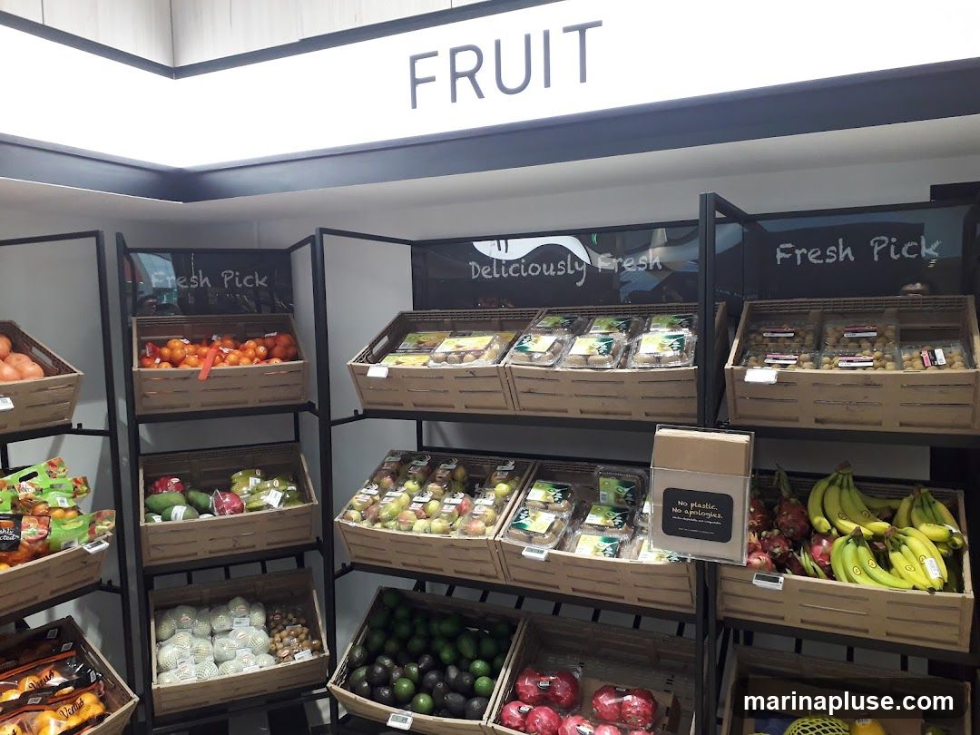 A display of various fruits in a store. The fruits are arranged in wooden crates on shelves. Above the display, a sign reads "FRUIT". There are also signs that say "Fresh Pick" and "Deliciously Fresh". Fruits visible include oranges, tomatoes, cucumbers, apples, bananas, dragon fruit, avocados, and pomegranates. Some fruits are packaged in plastic containers. A sign that says "No plastic. No apologies." is also visible.