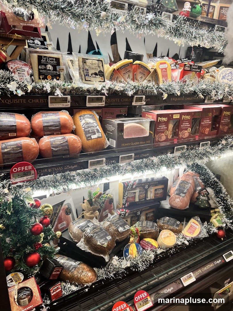 A refrigerated display case filled with various Christmas-themed food items, including honey baked ham, honey glazed ham DIY, and a selection of cheeses. The display is decorated with artificial snow, tinsel, and small Christmas trees. Several items have "OFFER" labels with prices.