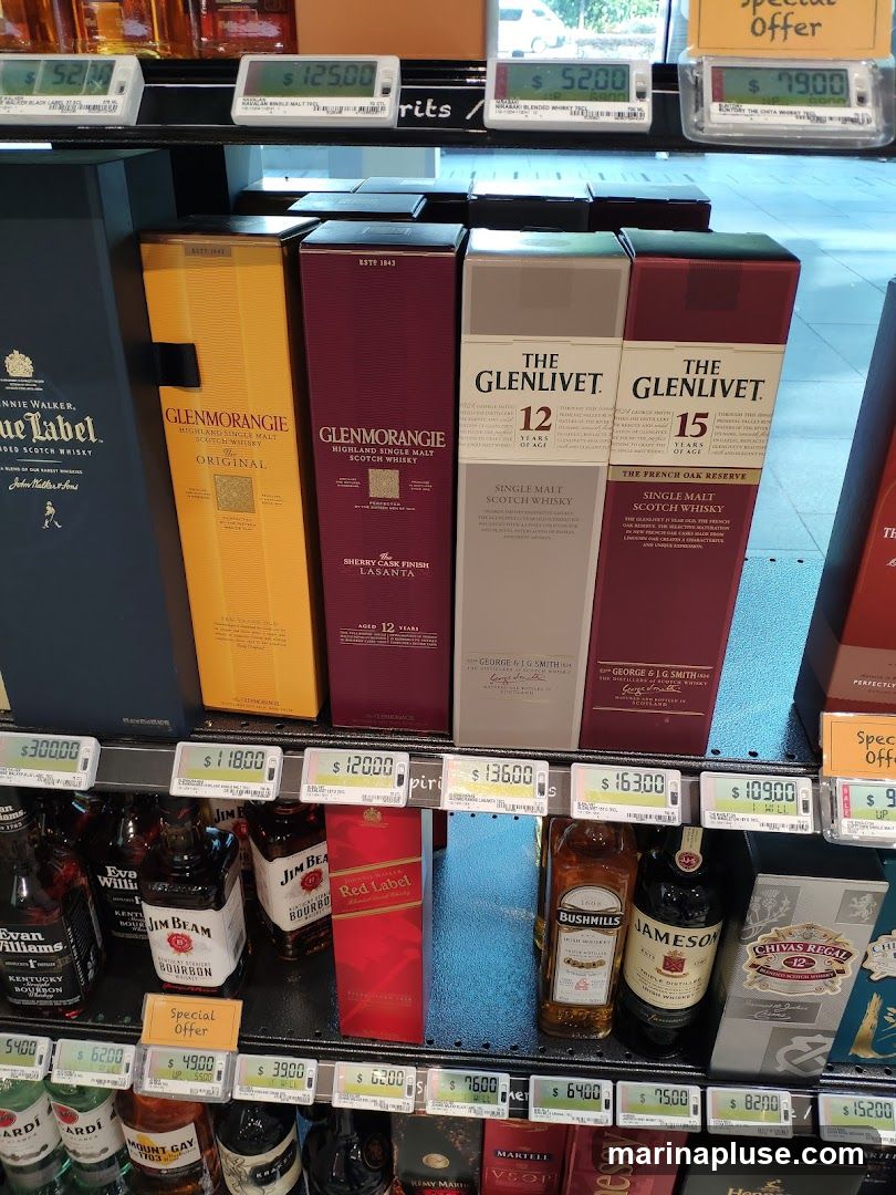 A retail display of various bottles and boxes of whiskey. Brands visible include Johnnie Walker, Glenmorangie, The Glenlivet, Evan Williams, Jim Beam, Bushmills, Jameson, and Chivas Regal. Prices are displayed digitally above the products.