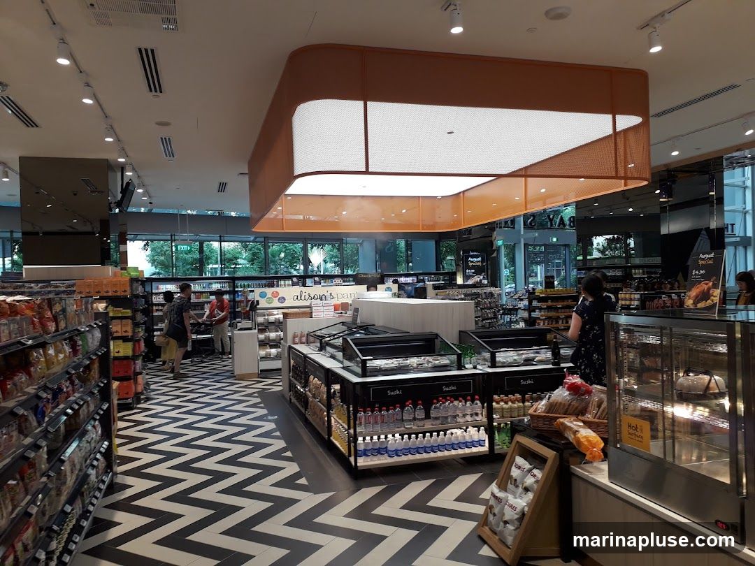 The image shows the interior of a brightly lit grocery store with aisles of products. The floor has a distinctive black and white zigzag pattern. In the center, there are refrigerated display cases and shelves stocked with various food items, including sushi and bottled drinks. A sign above a counter reads "alison's pantry". In the background, there are large windows offering a view of greenery outside. A promotional sign for an "August Special" featuring a roasted chicken is visible on the right.