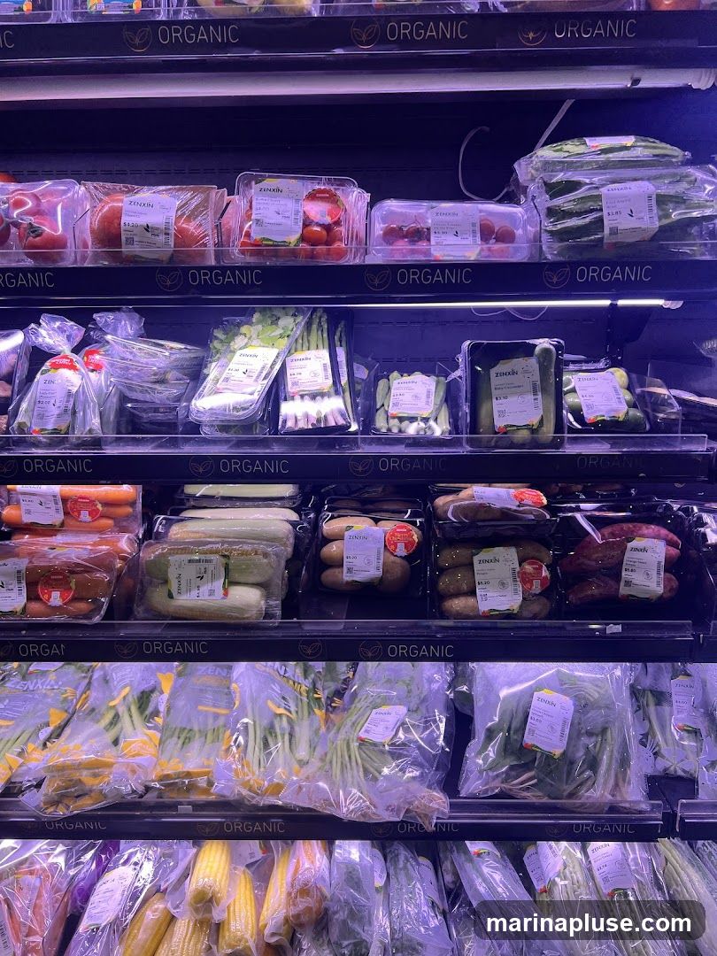 A refrigerated display case filled with various organic fruits and vegetables. The shelves are labeled "ORGANIC" and feature items such as tomatoes, cucumbers, asparagus, corn, and leafy greens, all packaged in plastic containers or bags.