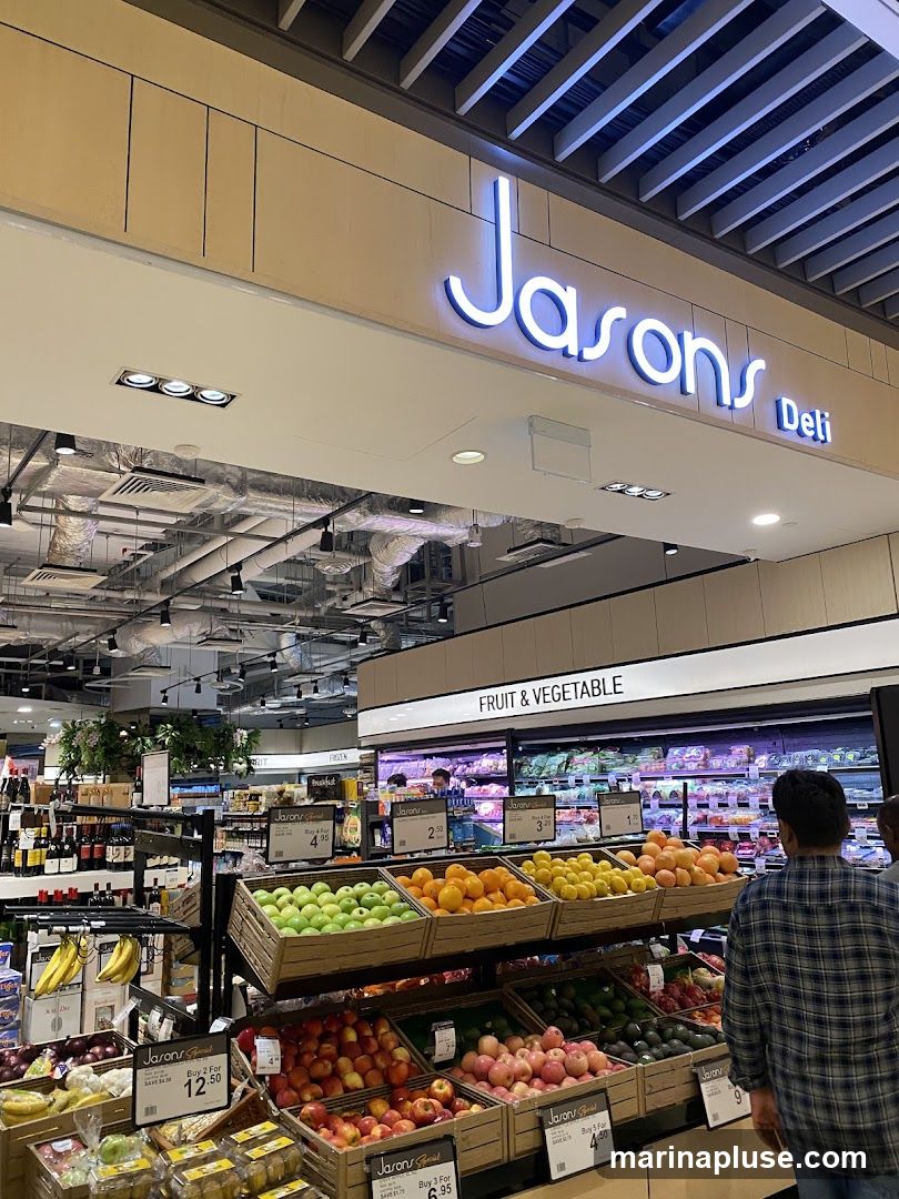 A view inside Jasons Deli, showing a well-stocked fruit and vegetable section. Displays of apples, oranges, lemons, and other produce are visible, with pricing signs. The store's signage "Jasons Deli" is prominently displayed above the entrance.