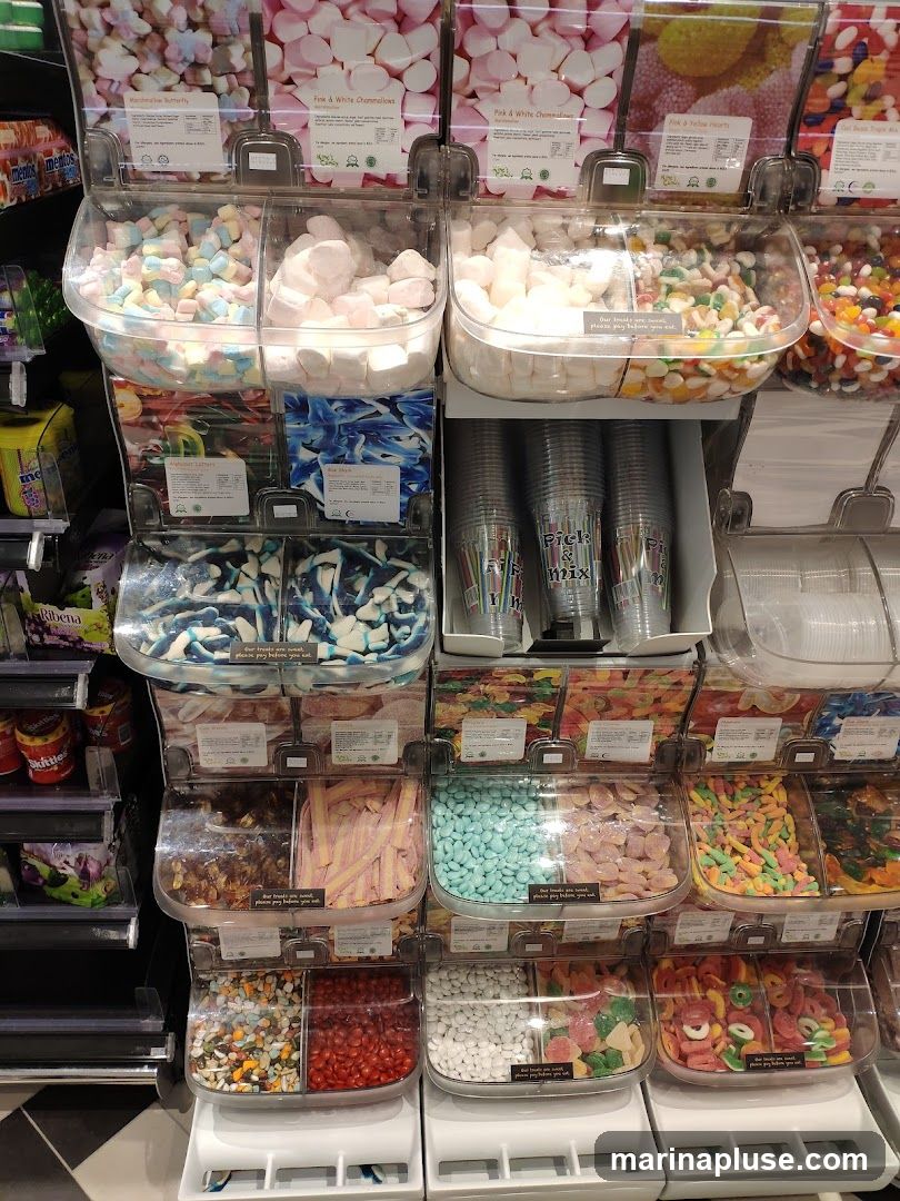 A wall of clear plastic bins filled with various types of candy. The bins are stacked in multiple rows and columns, showcasing a colorful assortment of sweets. Some bins contain marshmallows, gummy worms, jelly beans, and other confectionery items. Labels are visible above some of the bins, indicating the type of candy within. Cups for the pick-and-mix selection are also visible.