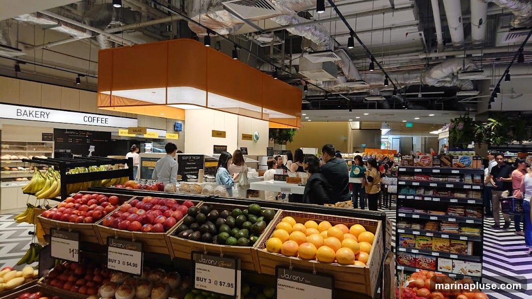 A wide shot of the interior of a grocery store, focusing on the produce section. Baskets of apples, avocados, and grapefruits are prominently displayed in the foreground. In the background, shelves are stocked with various snacks and other grocery items. Signs indicate "Bakery" and "Coffee" sections. Several shoppers are visible browsing the aisles.