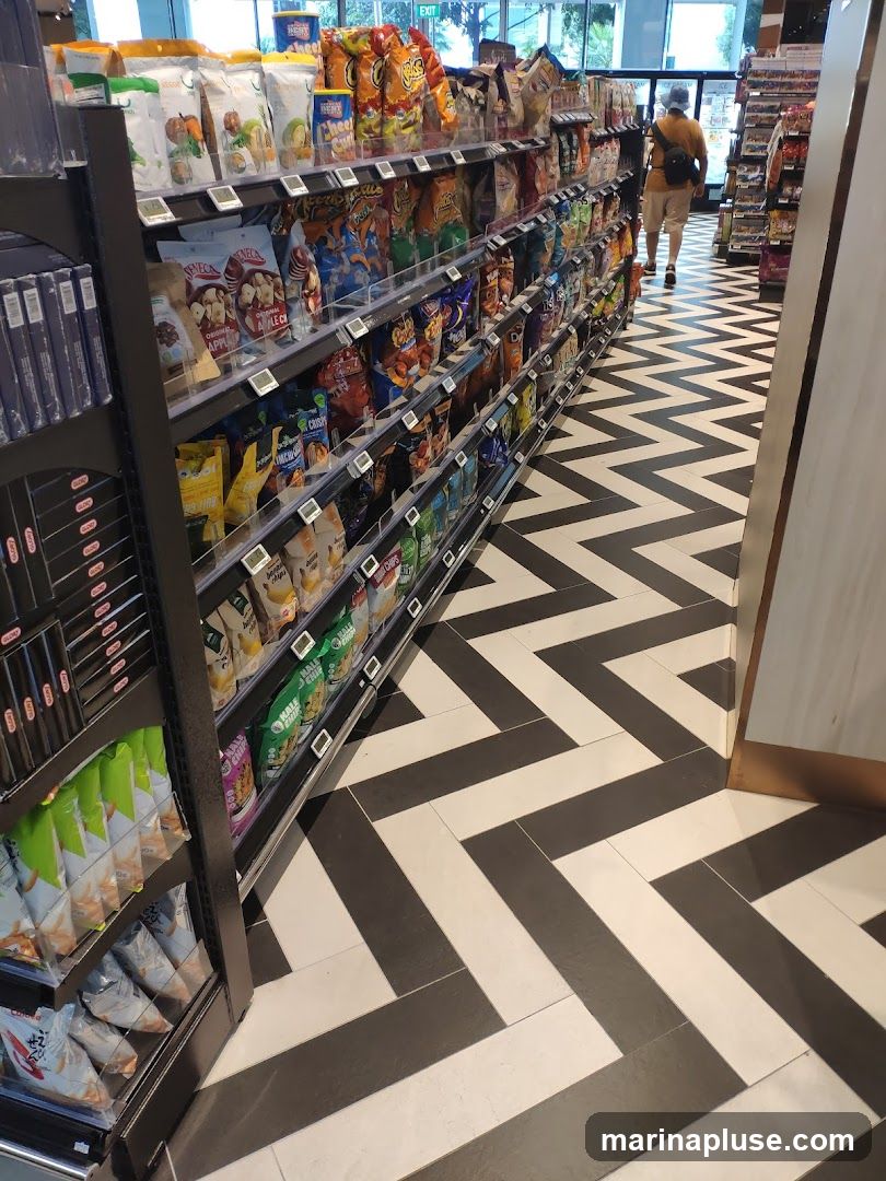 A long aisle in a store filled with shelves of snacks. The shelves are stocked with various bags of chips, nuts, and other packaged snacks. The floor has a distinctive black and white chevron pattern. A person is walking away from the camera down the aisle.
