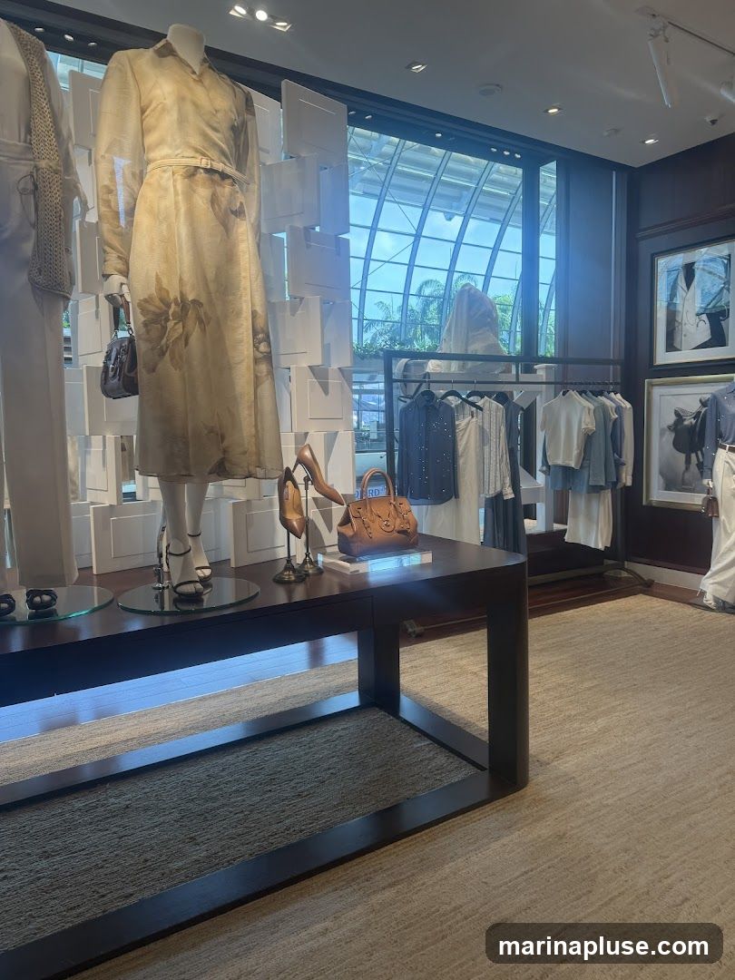 A well-lit clothing store interior with mannequins displaying dresses and racks of clothing. A dark wooden table in the foreground holds a handbag and shoes. The background features a large window with a view of greenery and a modern architectural structure. Framed pictures hang on the wall.