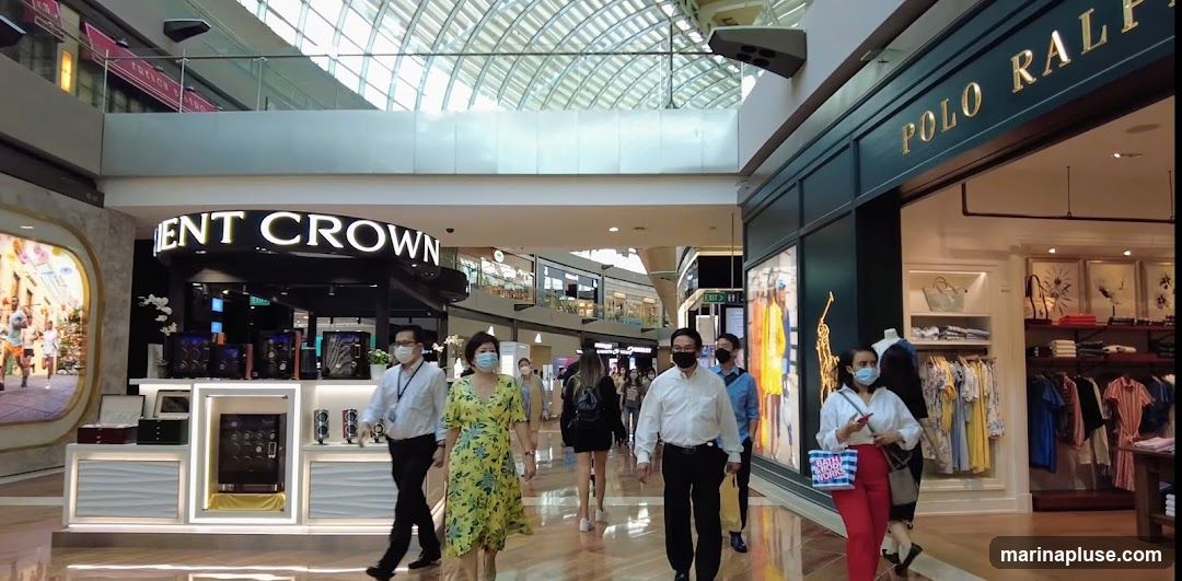 People walk through a brightly lit indoor mall. On the right, a Polo Ralph Lauren store displays clothing. On the left, a store with a sign reading "ENT CROWN" showcases watches and accessories.