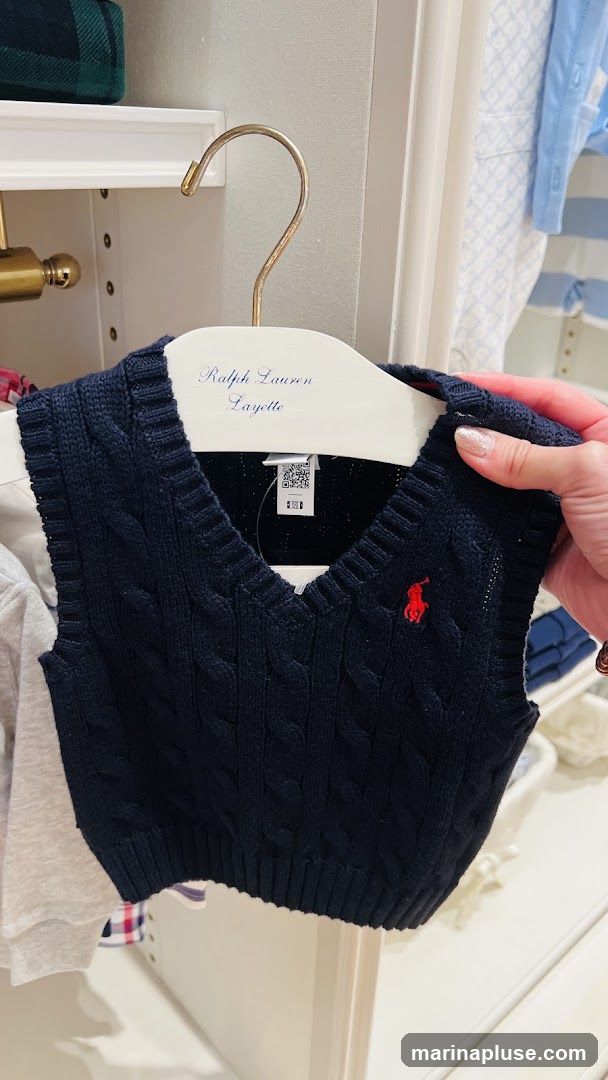 A navy cable knit vest with a red embroidered polo player logo is displayed on a white hanger. The hanger has "Ralph Lauren Layette" written in blue script. The vest is on display in a closet or store setting with other clothing items visible in the background.