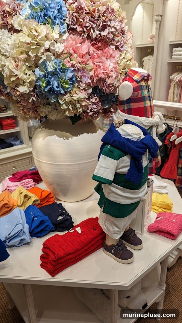 A display of children's clothing featuring a mannequin dressed in a polo shirt and sweater, surrounded by folded shirts and sweaters in various colors. A large vase filled with colorful hydrangeas is also part of the display. Shelves with more clothing are visible in the background.