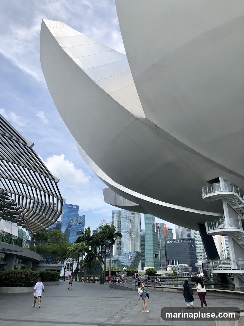 A wide shot of a modern architectural structure with a curved, white facade, set against a backdrop of a bustling cityscape. In the foreground, a paved plaza is populated by a few pedestrians. Palm trees and other greenery are interspersed between the buildings. The sky is partly cloudy.