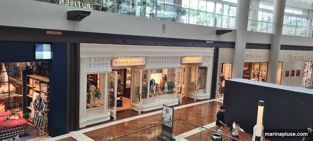 The exterior of a Ralph Lauren store is visible, featuring a classic white facade with large display windows. Mannequins dressed in clothing are displayed inside. The store's name, "RALPH LAUREN," is prominently displayed on a sign above the entrance. Adjacent to the Ralph Lauren store, a Dolce & Gabbana store is also visible.
