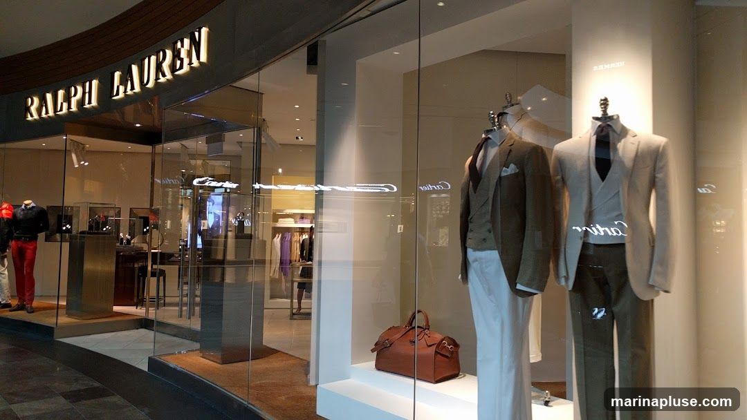 The exterior of a Ralph Lauren store is visible, with a large glass window displaying mannequins dressed in suits and a brown leather duffel bag. The Ralph Lauren logo is illuminated above the entrance. Inside the store, clothing racks with various garments and display cases with accessories can be seen.