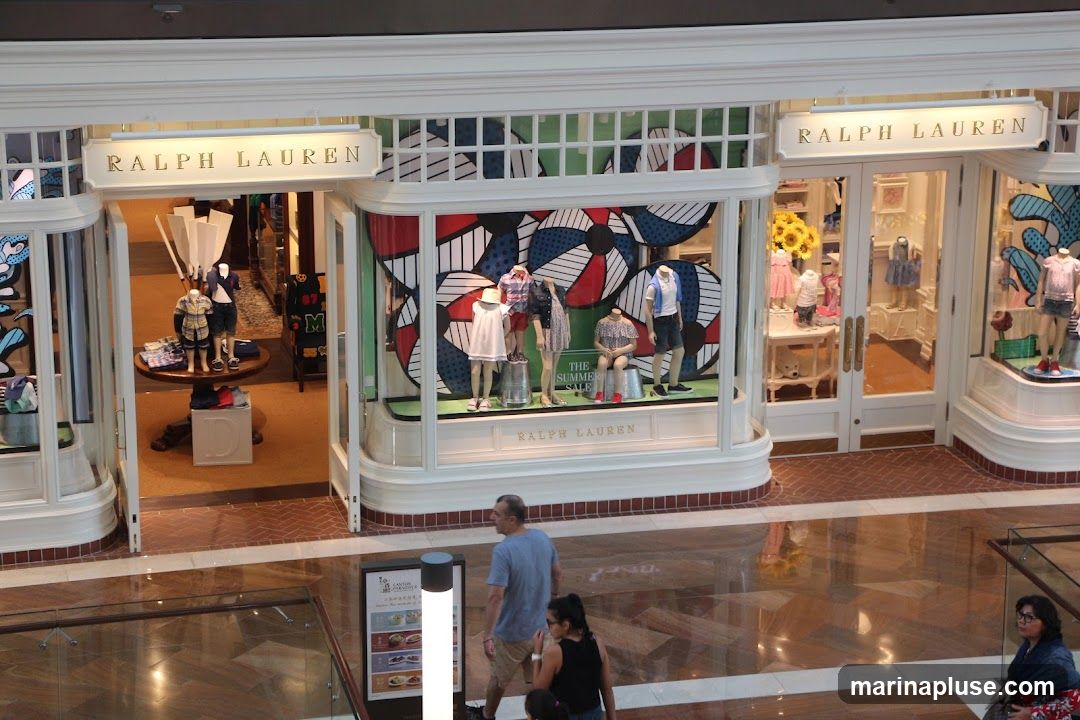 The exterior of a Ralph Lauren store with mannequins displaying clothing in the windows. The store has large glass windows and double doors. The Ralph Lauren logo is visible above the entrance and on the window displays.