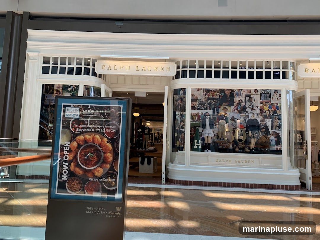 A storefront for Ralph Lauren with mannequins in the window display. A large advertisement for a restaurant called Justin is placed in front of the store. The advertisement features images of food and text in English and Chinese. The Ralph Lauren signage is visible above the entrance and on the window display.