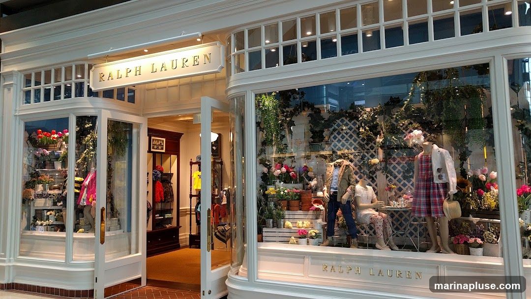 The exterior of a Ralph Lauren store with a white facade. The store features large display windows showcasing mannequins dressed in clothing, surrounded by floral arrangements and greenery. The store's name, "RALPH LAUREN," is visible in gold lettering above the entrance and on the lower part of the display window. An open door reveals a glimpse of the store's interior, with clothing racks and displays.