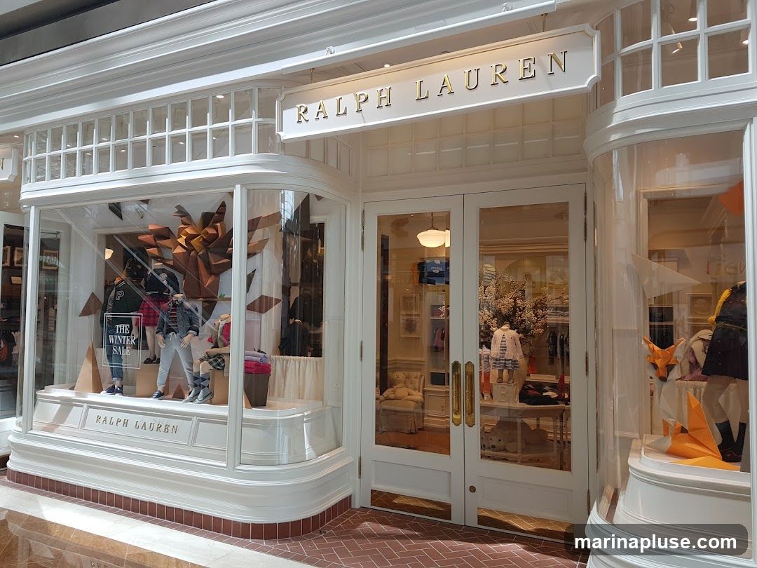 The exterior of a Ralph Lauren store with large display windows and double doors. The store name is prominently displayed above the entrance and on a ledge below the window. The windows showcase mannequins dressed in winter clothing and decorative elements.