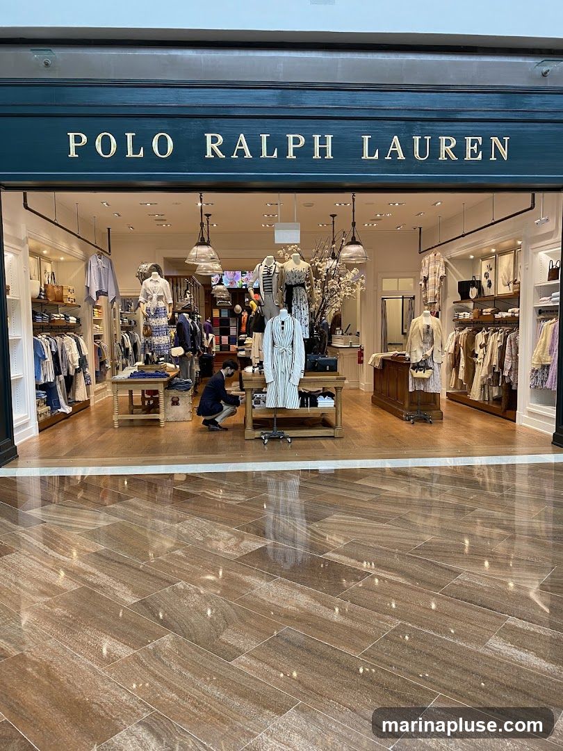 The exterior of a Polo Ralph Lauren store with the brand name displayed prominently above the entrance. The store is well-lit and showcases clothing on mannequins and racks. A person is seen inside adjusting a display. The floor in front of the store has a polished, wood-like tile pattern.