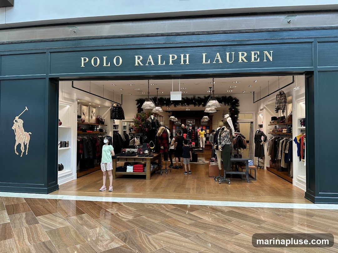 The exterior of a Polo Ralph Lauren store with the brand's logo and name prominently displayed. Inside, clothing is displayed on racks and tables, with mannequins showcasing outfits. Several people are browsing the merchandise.
