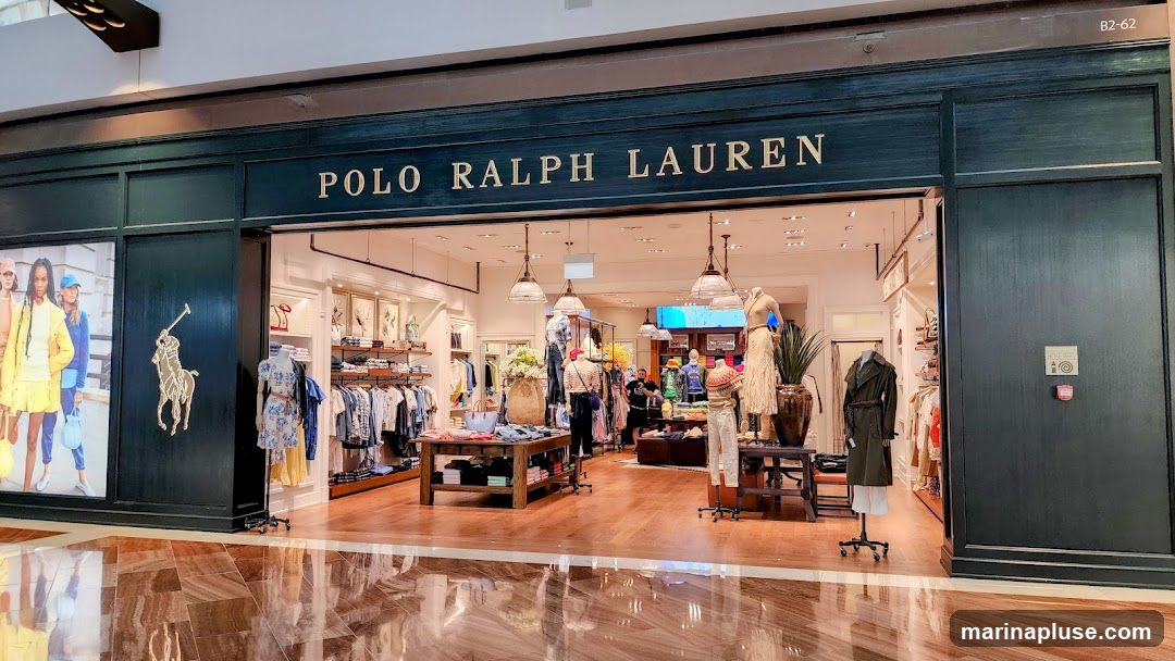 The exterior of a Polo Ralph Lauren store. The store features a dark green facade with the brand name "POLO RALPH LAUREN" in gold lettering above the entrance. A large gold polo player logo is visible on the left side of the storefront. Inside, clothing racks and display tables showcase various apparel items. Mannequins are dressed in outfits, and the store appears well-lit and organized.