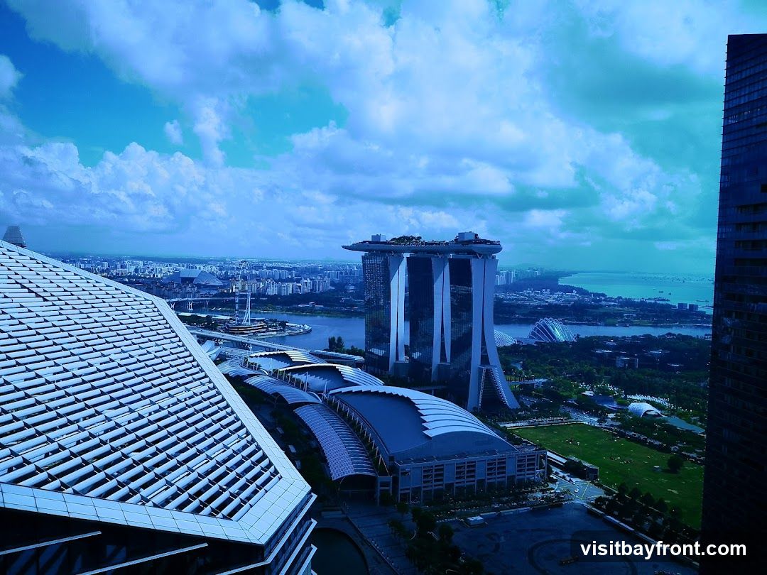 A panoramic view of the Singapore skyline, featuring the iconic Marina Bay Sands hotel, Gardens by the Bay, and other modern architecture under a cloudy blue sky. The foreground shows the distinctive roof of a building.