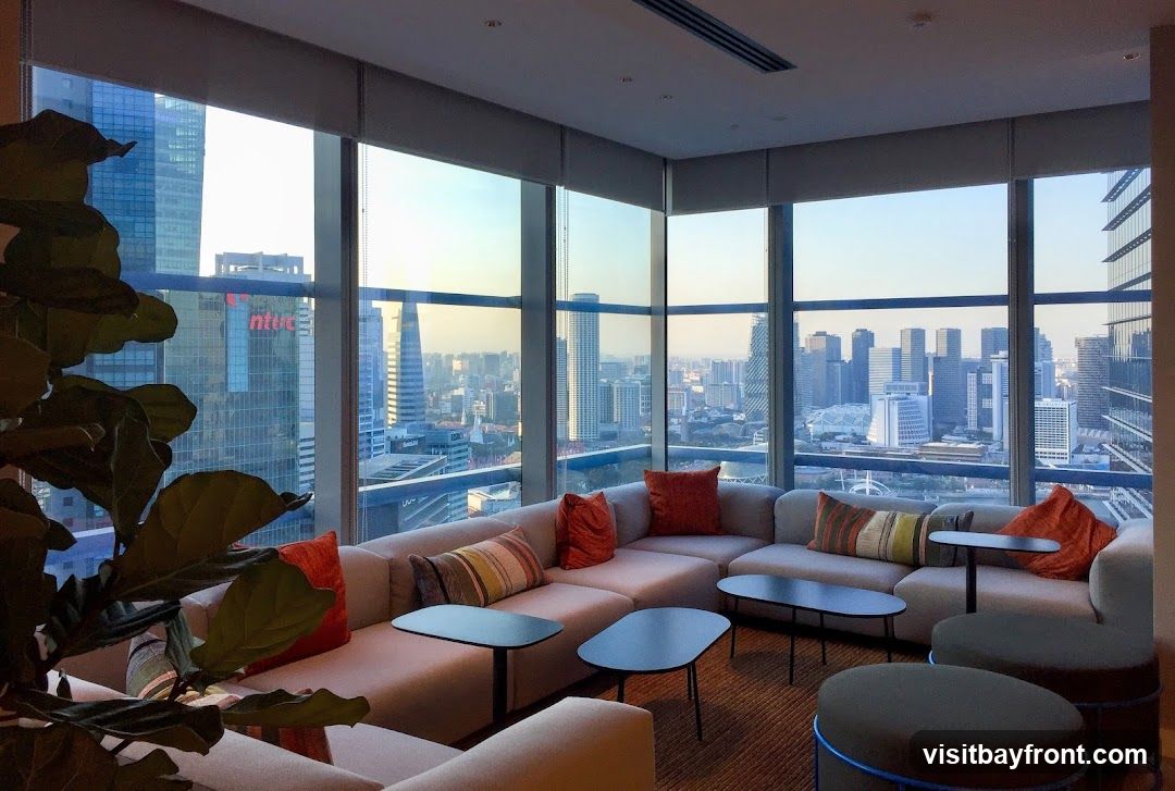 A contemporary lounge area with a large sectional sofa, accent pillows, and low coffee tables, offering panoramic views of a bustling cityscape at sunset. A large potted plant is visible in the foreground.
