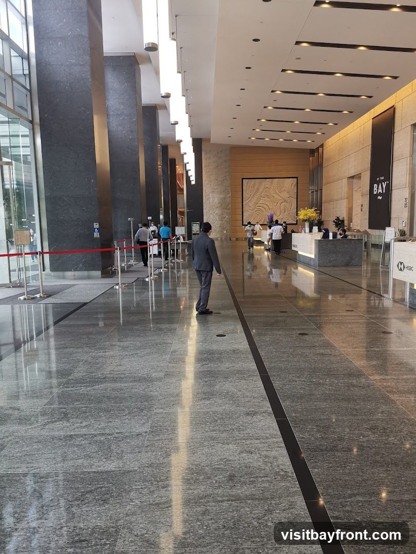 A wide, polished marble floor stretches through a contemporary office building lobby. Tall, dark marble-clad columns line the left side, with a glass facade revealing glimpses of the outside. Modern, elongated light fixtures hang from the high ceiling. In the distance, a reception desk is visible with staff attending to visitors. A large, textured art piece adorns the wall behind the reception area. A black banner with white text reading "BY THE BAY" is displayed on the right side, along with an HSBC logo. Several people are walking through the lobby, some in business attire. Red stanchions with ropes guide a queue near the entrance on the left.