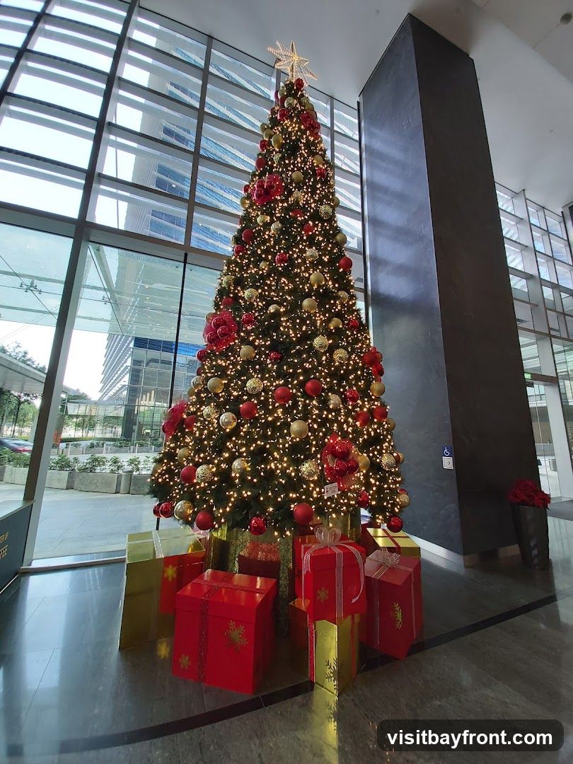A tall, decorated Christmas tree stands in a modern lobby, adorned with lights, red and gold ornaments, and topped with a star. Several wrapped gift boxes in red and gold are placed around its base. The background features large windows with a view of office buildings and a dark, textured pillar.