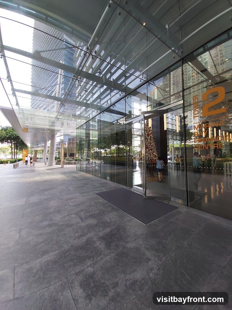A modern glass-fronted building entrance with a covered walkway. The building has large glass panels and a prominent orange number '2' on the glass, along with text that reads 'MARINA BAY FINANCIAL CENTRE'. People are seen walking in and out of the building. A Christmas tree is visible inside the lobby.