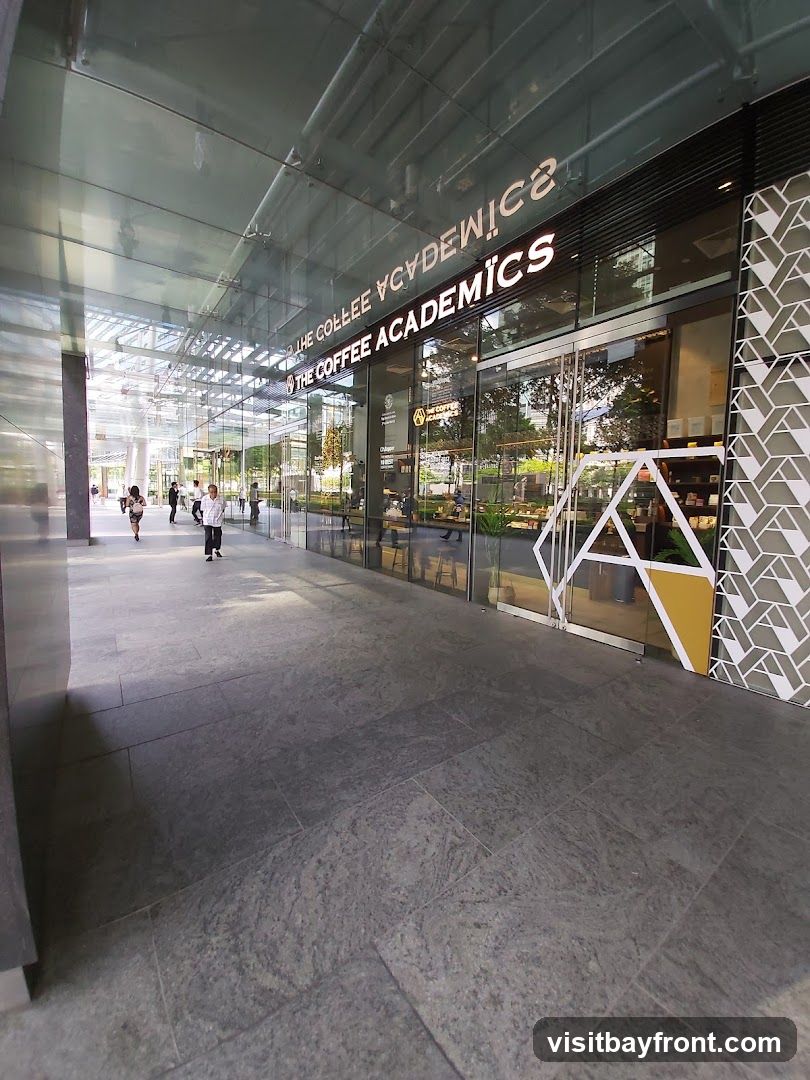 A wide shot of the exterior of The Coffee Academics cafe. The storefront features large glass windows and doors, with the cafe's name prominently displayed in white lettering above the entrance. The interior is visible through the windows, showing seating areas and displays. The facade is decorated with a geometric pattern on the right side. People are walking by on the tiled walkway in front of the cafe.