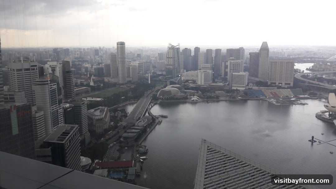 An aerial view of a dense cityscape with numerous skyscrapers and a large body of water in the foreground. The sky is overcast. Several distinct buildings, including a boat-shaped structure and a tall, slender tower, are visible. Roads and bridges crisscross the urban landscape.