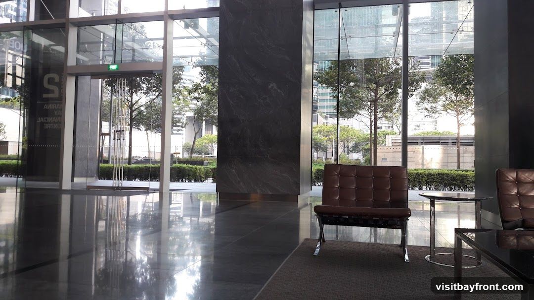 A spacious and modern lobby with a polished floor reflecting the natural light. The entrance area features glass doors and windows, with a large marble-clad pillar. Comfortable brown leather armchairs and small tables are arranged for waiting guests. Outside, trees and modern buildings are visible through the glass facade.