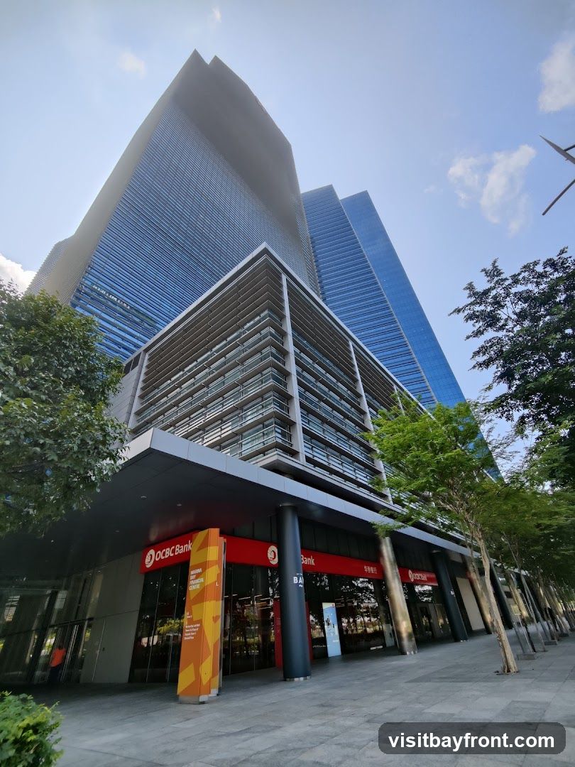 A low-angle shot of a modern bank building with a glass facade and a prominent red and white OCBC Bank sign. Tall, sleek skyscrapers rise behind it, and trees line the sidewalk in front.