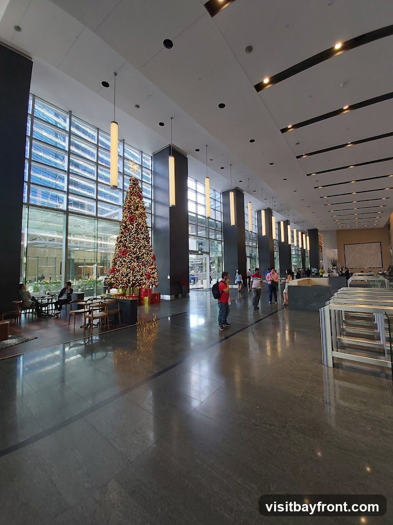 A spacious and modern lobby with a tall Christmas tree decorated with ornaments and lights. The lobby features a polished dark floor, large windows with a view of a cityscape, and contemporary lighting fixtures. Several people are seen walking through the lobby or sitting at tables.