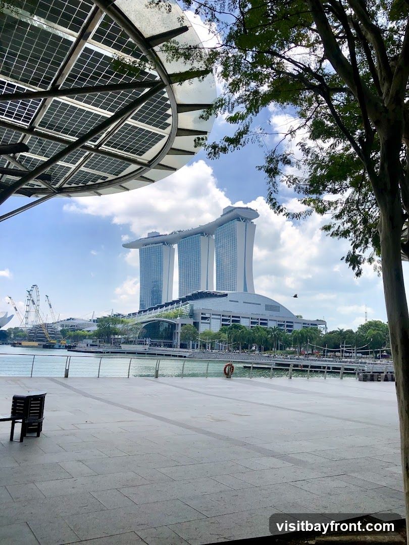 A view of the Marina Bay Sands complex in Singapore, with its distinctive three towers and sky park. The foreground shows a paved walkway with a few chairs, and trees partially obscure the view. The sky is partly cloudy.