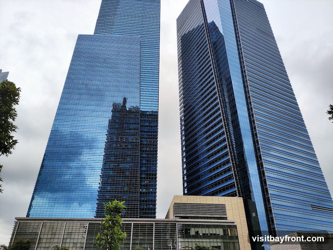 A low-angle shot of two modern skyscrapers with blue glass facades against a cloudy sky. The buildings have a sleek, contemporary design with many windows. Trees are visible in the foreground and on the sides, adding a touch of nature to the urban landscape.