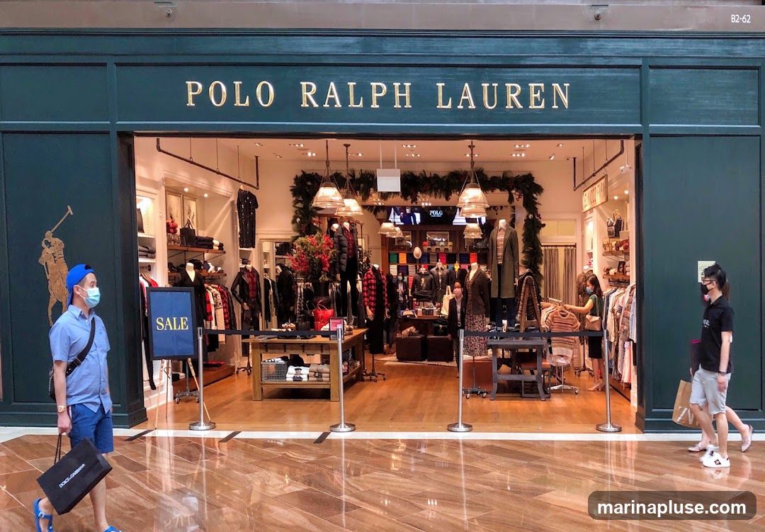 The exterior of a Polo Ralph Lauren store. The store has a dark green facade with gold lettering that reads "POLO RALPH LAUREN" above the entrance. A large Polo player logo is visible on the left side of the facade. Inside, clothing is displayed on racks and shelves, and mannequins are dressed in various outfits. Several people are seen inside and outside the store, some wearing masks. A blue sign with the word "SALE" is placed near the entrance.