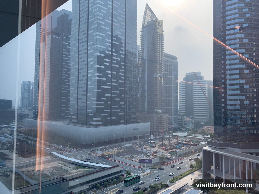 A panoramic view of a modern cityscape dominated by tall skyscrapers. In the foreground, a construction site is visible with cranes and heavy machinery. Roads with cars and a bus are present, indicating urban activity. The sky is hazy, suggesting either smog or a cloudy day. Reflections on the glass from which the photo is taken are also visible.