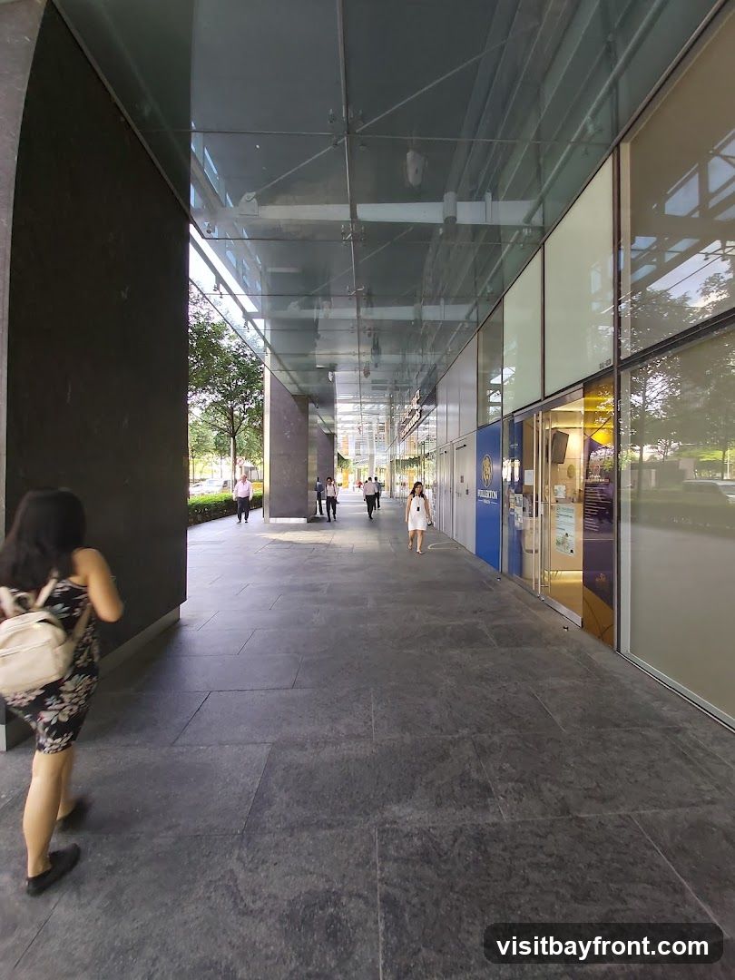 A covered walkway leads to a row of storefronts. A woman with a backpack walks away from the camera on the left. Several people are walking towards the storefronts in the distance. The storefronts have large glass windows and doors, with some signage visible.