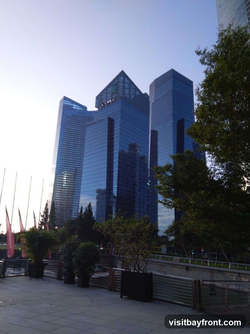 A view of several modern skyscrapers with glass facades against a clear sky. In the foreground, there is a paved walkway with planters containing trees and shrubs. Red flags are visible in the distance on the left. The scene appears to be an urban waterfront area.