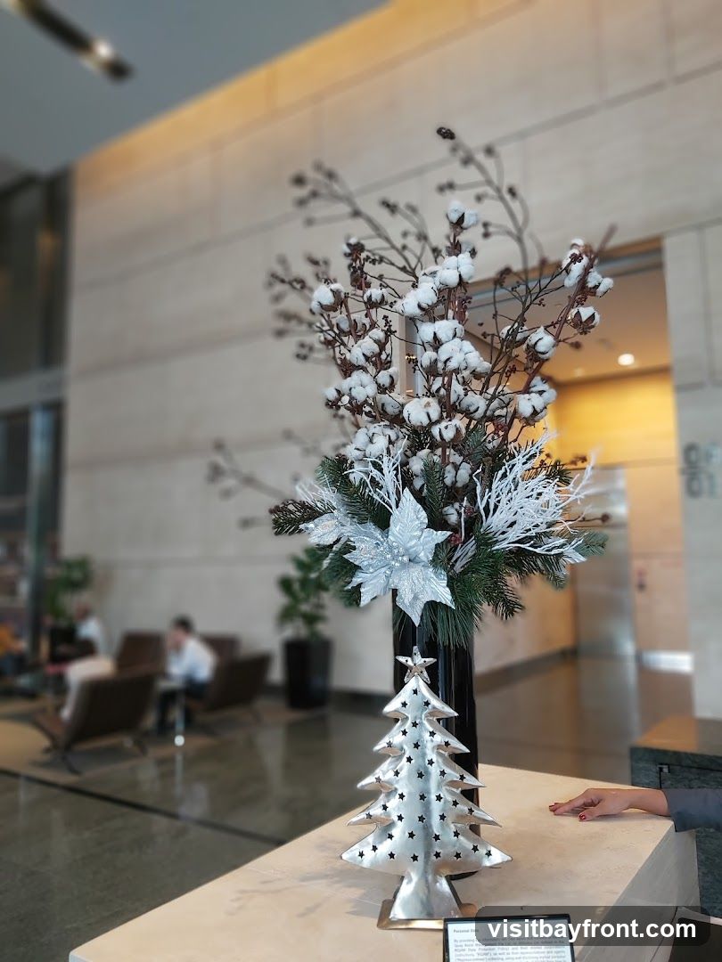 A festive holiday decoration featuring a silver Christmas tree and a floral arrangement with cotton branches and silver poinsettias is displayed on a counter. In the background, a blurred view of a lobby or office space with seating and a doorway is visible.