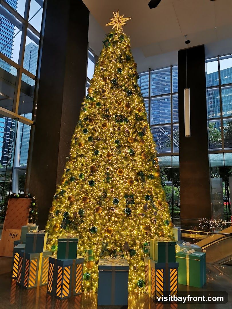 A large, brightly lit Christmas tree decorated with gold and green ornaments stands in a lobby. The tree is topped with a star. Around the base of the tree are several decorative gift boxes. In the background, large windows reveal modern buildings.