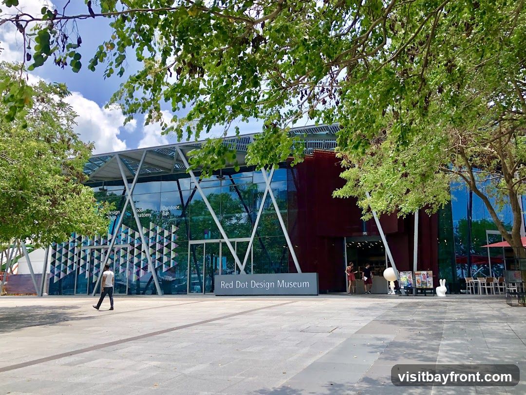The Red Dot Design Museum is a modern building with a glass facade and a dark red section. A sign with the museum's name is visible in front of the building. Trees provide shade in the foreground.