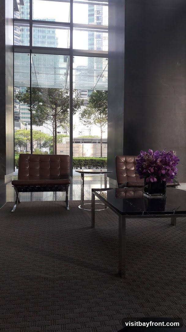 A contemporary lobby seating area features two brown leather Barcelona chairs and a black marble-topped coffee table with a vase of purple flowers. The space is carpeted and has large windows offering a view of city buildings and trees.