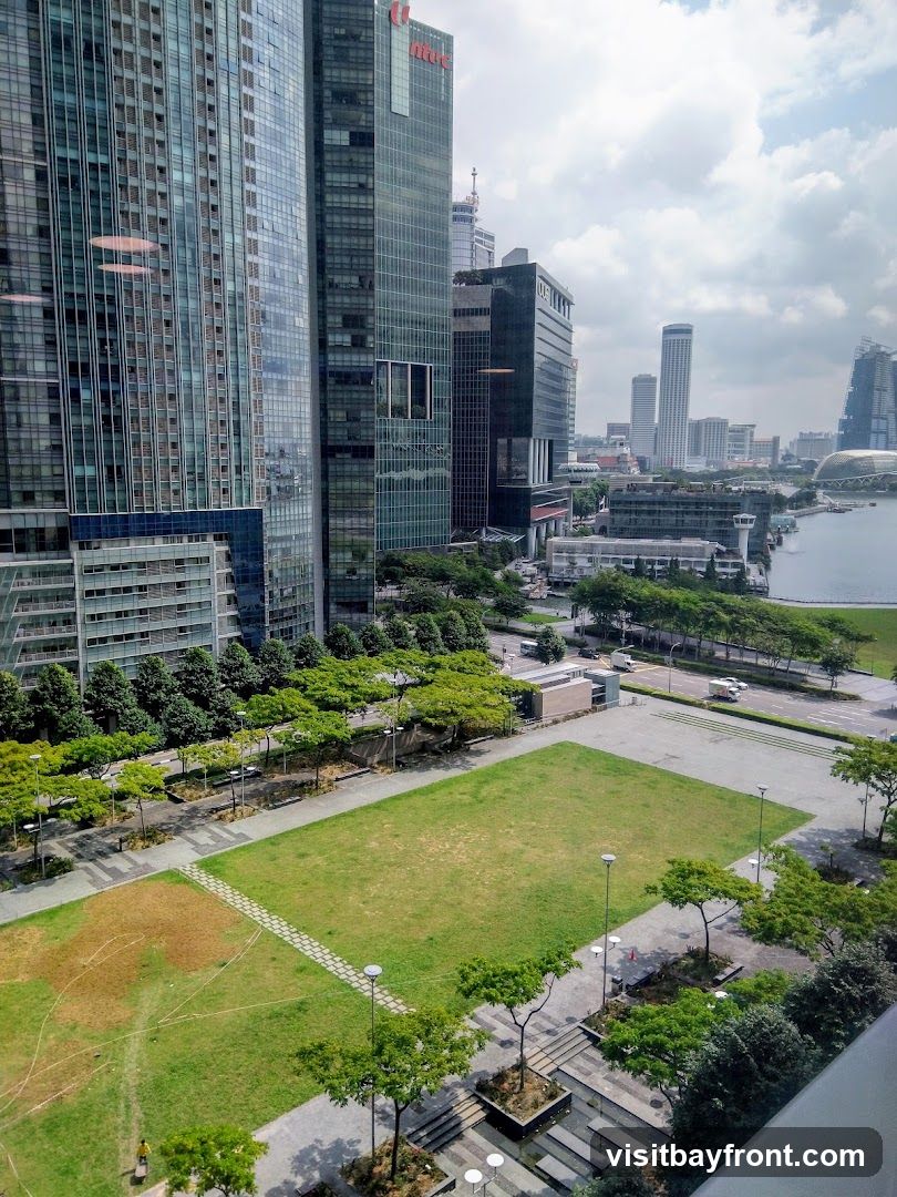 A high-angle view of a modern cityscape featuring numerous tall glass skyscrapers and a large green park area in the foreground. A body of water is visible in the distance, with more buildings lining the waterfront. The sky is partly cloudy.