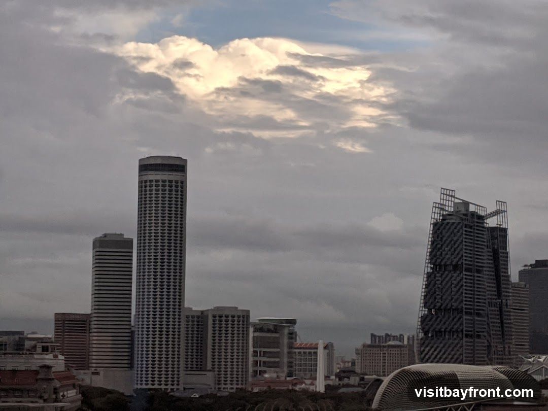 A panoramic view of a city skyline featuring several tall skyscrapers under a dramatic, cloudy sky. The clouds are illuminated from above, creating a contrast with the darker, overcast sky. In the foreground, the tops of some buildings and structures are visible, suggesting a dense urban environment.